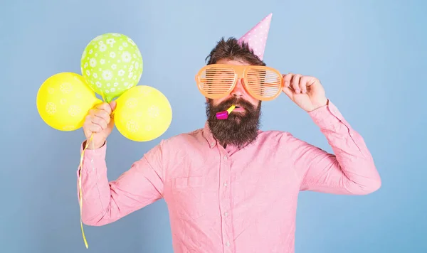 Hipster mit buschigem Bart feiert Geburtstag. Bärtiger Mann posiert in Geburtstagskappe mit riesiger Brille und bunten Luftballons auf blauem Hintergrund. Mann mit gestutztem Bart bläst Party-Pfeife — Stockfoto