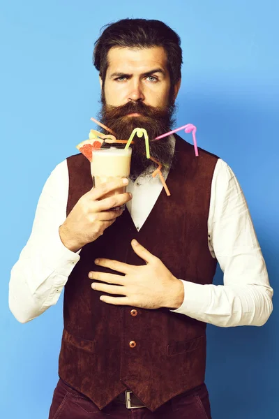 Homem barbudo bonito com tubos coloridos em barba longa e bigode tem cabelo elegante no rosto sério segurando vidro de coquetel alcoólico na cintura de couro de camurça vintage no fundo do estúdio azul — Fotografia de Stock