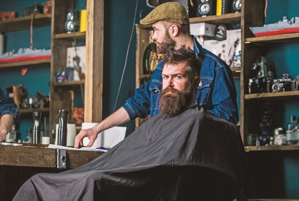 Man with beard covered with black cape waiting while barber changing clipper grade. Hipster client getting haircut. Client with beard ready for trimming or grooming. Haircut process concept