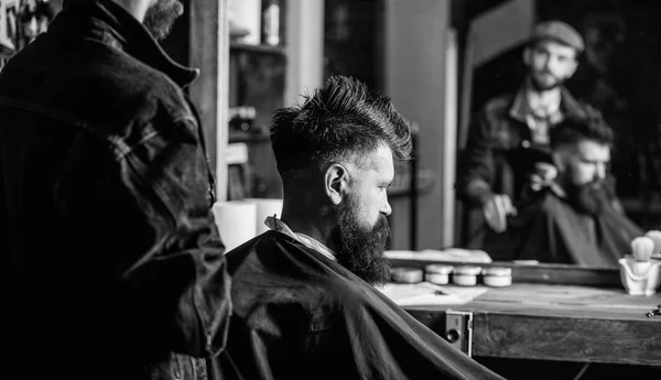 Concept de salon de coiffure. Homme avec barbe recouverte de cape noire se trouve dans la chaise de coiffeur, fond miroir. Hipster avec barbe attend le coiffeur et la coupe de cheveux. Homme avec barbe client du salon de coiffure hipster — Photo