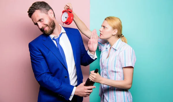 After corporate party. Man suffering from alcoholism. Angry wife meeting drunk husband late at home. Businessman with alcohol bottle and woman with alarm clock. Addictive alcoholism or alcohol abuse — Stock Photo, Image