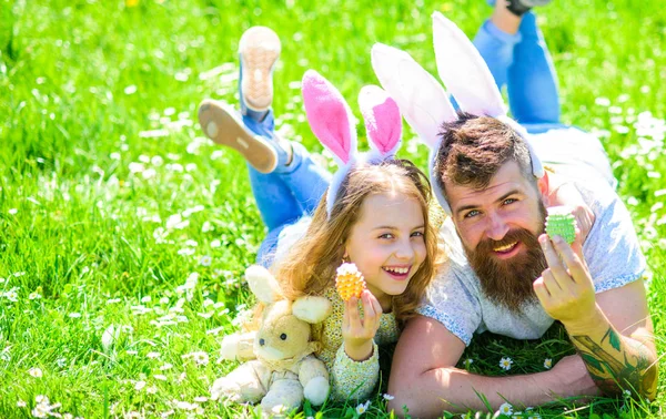 Koncepce rodinné dovolené. Dítěte a muž s roztomilý zajíček uši hrát společně. Dívka s tátou ležící na trávě na slunečný den. Otec a dcera na šťastné tváře pořádá velikonoční vajíčka, tráva na pozadí — Stock fotografie
