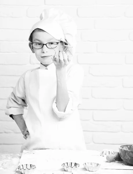 Młody chłopak małych cute kucharz kucharz w biały uniform i kapelusz na twarz Witraż mąki z okularami stojący w pobliżu tabeli z wałkiem czerwony bowl i przytrzymując cookie cutter star na tle ściany Cegła — Zdjęcie stockowe