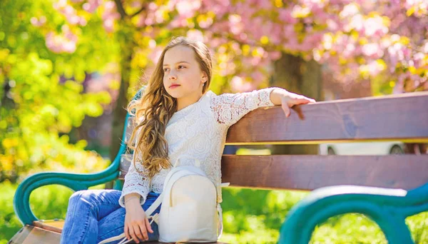 Menina no rosto sorridente senta-se no banco, sakura no fundo, desfocado. Menina relaxante enquanto caminhe no parque perto de flor de cereja. Criança bonito com mochila desfrutar de dia de primavera ensolarado. Conceito de acessórios de moda — Fotografia de Stock