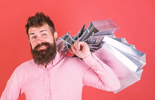 Hombre con barba y bigote lleva bolsas de compras en el hombro, fondo rojo. Hombre de compras en temporada de ventas con descuentos. Concepto de compras. Hipster en la cara feliz es adicto a las compras o adicto a las compras —  Fotos de Stock