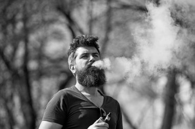 Elektronik Sigara kavramı. Uzun sakallı ve bulutlar duman görünüyor rahat olan adam. Sakallı adam vape güneşli günde sigara içiyor. Adam sakal ve bıyık sakin yüzündeki defocused arka plan, dallarda