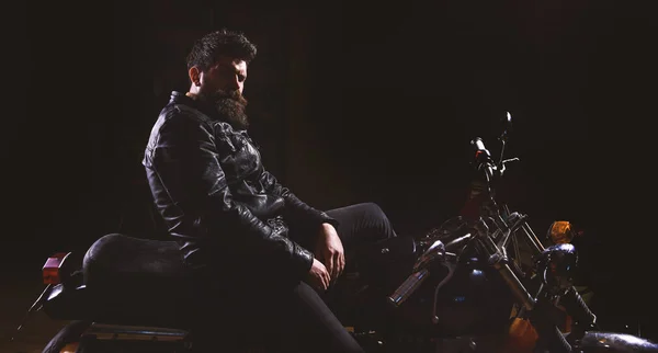 Hombre con barba, motorista con chaqueta de cuero sentado en moto en la oscuridad, fondo negro. Macho, motero brutal en chaqueta de cuero montando motocicleta por la noche, espacio para copiar. Concepto de corredor nocturno — Foto de Stock