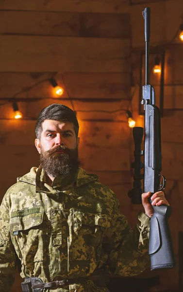 Gamekeeper concept. Hunter, brutal hipster with gun in his hand ready for hunting. Macho on strict face at gamekeepers house. Man with beard wears camouflage clothing in wooden interior background — Stock Photo, Image