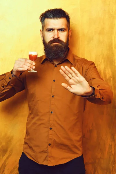 Bearded man, long beard. Brutal caucasian serious hipster with moustache in brown shirt holding alcoholic red shot and showing stop on golden texture backgroun