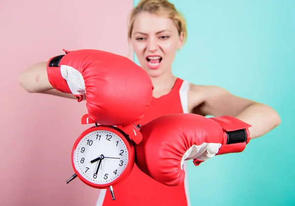 Its time for boxing training. Emotional woman holding clock in boxing gloves. Cute boxer with angry look fighting with alarm clock. Sportswoman punching a clock. There is still time on the clock