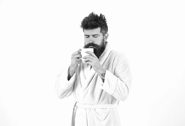 Macho drowsy, sleepy face drinks coffee in morning enjoying aroma. Morning rituals concept. Man with beard and disheveled hair stands in bathrobe, holds mug with tea or coffee, white background