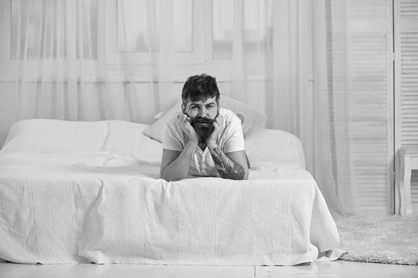 Chico en la cara tranquila acostado en el borde de la cama en sábanas blancas. Hombre acostado en la cama, cortinas blancas en el fondo. Concepto de dormitorio y colchón. Macho con barba y bigote relajante, descansando . —  Fotos de Stock