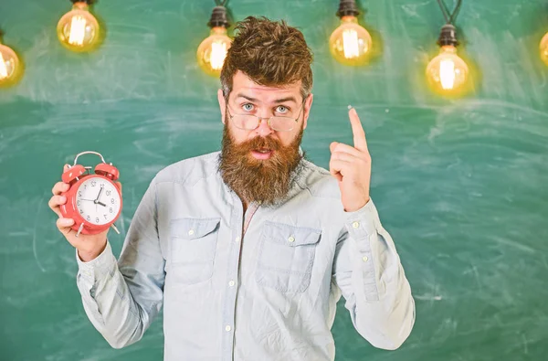 El profesor de anteojos tiene despertador. El hipster barbudo sostiene el reloj, advirtiendo con el dedo índice, copiando espacio. Concepto de disciplina. Hombre con barba y bigote en la cara estricta de pie en el aula —  Fotos de Stock