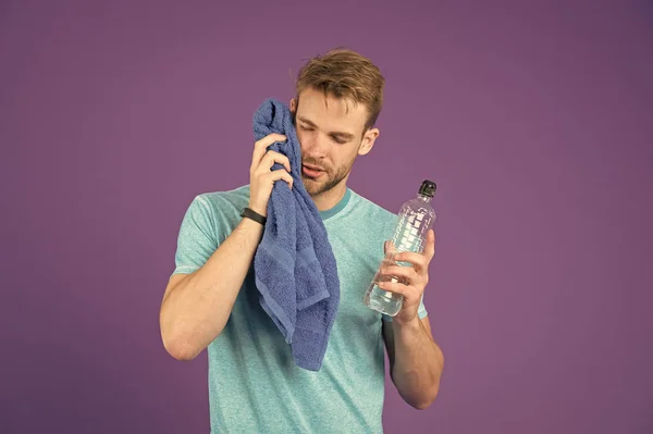 Atleta en camiseta azul y pantalones cortos con toalla después del entrenamiento. El hombre sostiene la botella de agua sobre fondo violeta. Deportista con botella de plástico. Sed y deshidratación. Actividad deportiva y energía —  Fotos de Stock