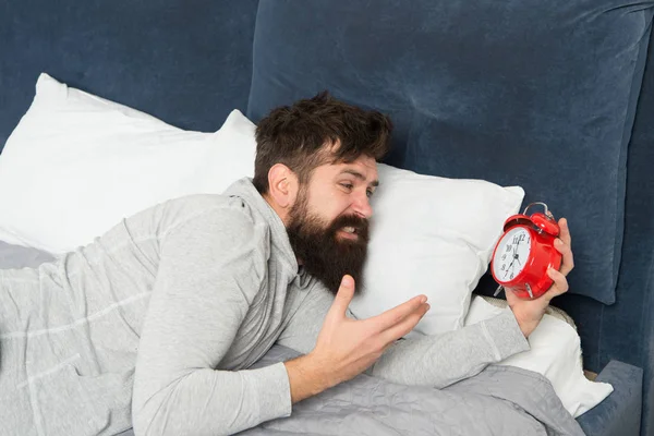 Túl korán van. A szakállas ember utálja az ébresztőórát, miközben otthon fekszik az ágyban. férfi ébresztőórával. Ébredj! Az ember az ágyban alszik. Ideje szünetet tartani. Depressziós ember. Megint elkéstél. Fáradtság és stressz — Stock Fotó