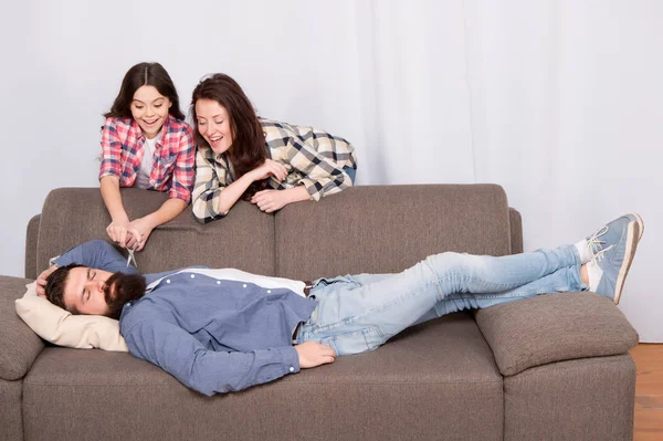 Lelucon keluarga. Gadis kecil dengan jenggot ibunya. Keluarga bahagia di rumah. Pria berjanggut santai di sofa. Wanita dengan anak perempuan. Memotong jenggot ayah anak. Jenggot memotong lelucon. Jenggot — Stok Foto