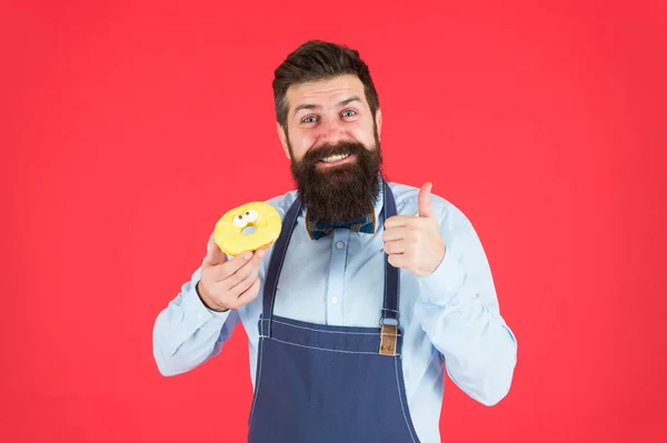 Conceito de café e padaria. Doce donut do padeiro. Homem padeiro barbudo no avental de cozinha segurar sobremesa bonito. Formas de reduzir a fome e o apetite. Hipster barbudo padeiro segurar donut envidraçado no fundo vermelho — Fotografia de Stock