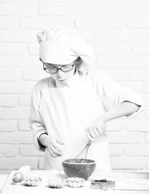 genç çocuk küçük sevimli aşçı Şef beyaz üniforma ve tablo ile oklava kalıpları yakınındaki kek kırmızı kase için ayakta ve tuğla duvar arka plan üzerinde pişirme gözlüklü mutlu yüz şapka