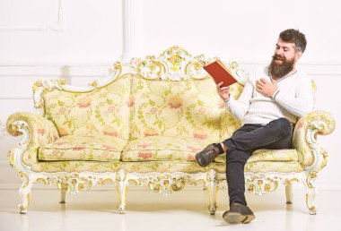 Adam zevk ile eski kitap okuma. Sakal ve bıyık oturur barok tarzı kanepe, tutar kitap, beyaz duvar arka plan üzerinde olan adam. Mizahi edebiyat kavramı. Maço yüz kitap okuma gülüyor üzerinde