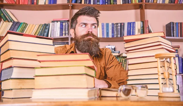 Homem no rosto estrito sentar entre pilhas de livros, enquanto estuda na biblioteca, estantes de livros no fundo. Conceito de bicho-de-biblioteca. Professor ou estudante com barba sente-se à mesa com ampulheta e óculos, desfocado — Fotografia de Stock