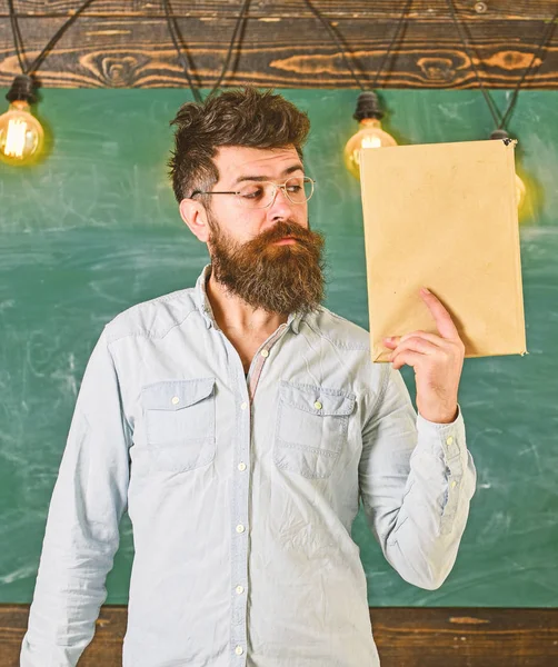 Profesor en anteojos tiene libro en la mano, espacio de copia. El hombre con barba y bigote en la cara tranquila se para en el aula. Científico sostiene libro, pizarra en el fondo. Concepto de literatura científica —  Fotos de Stock