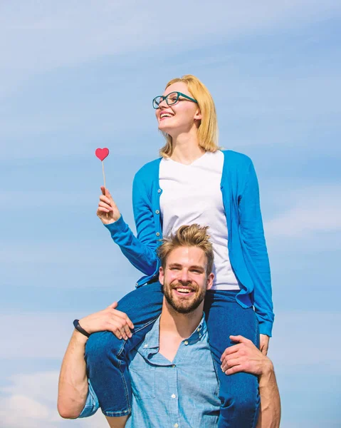 El hombre lleva a su novia en hombros, fondo del cielo. Pareja feliz cita divirtiéndose juntos. La mujer sostiene el corazón en el palo símbolo del amor. Concepto de fecha romántica. Pareja enamorada caminando al aire libre día soleado —  Fotos de Stock