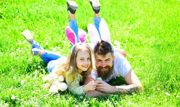 Concepto de trabajo en equipo. Papá y la niña encontraron huevos de Pascua en el juego de caza tradicional en. Hombre con barba y lindo niño yacía en la hierba con huevo de Pascua en la mano. Padre e hijo con orejas de conejo en el prado — Foto de Stock