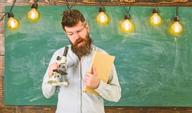 Gözlük öğretmen kitap ve mikroskop tutar. Sakal ve bıyık meşgul yüzünde olan adam. Bilimsel araştırma kavramı. Bilim adamı kitap ve mikroskop, kara tahta arka plan üzerinde tutar