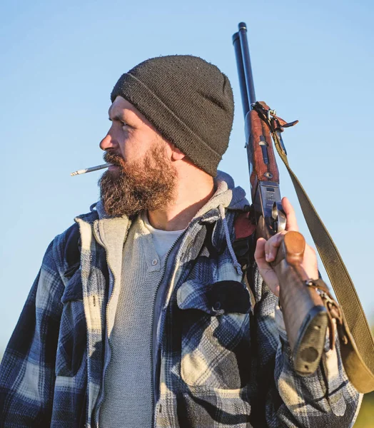 Caçando conceito de hobby masculino. Homem brutal barbudo cara gamekeeper azul céu fundo. Caçador com espingarda de perto. Guy caçador barbudo passar a caça ao lazer e fumar. Brutalidade e masculinidade — Fotografia de Stock