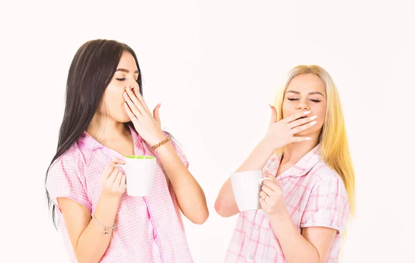 Meninas bebendo chá ou café de manhã, isolado em fundo branco. Loira e morena em rostos sonolentos bocejando, segura canecas com café. Irmãs ou melhores amigas de pijama. Conceito de café da manhã — Fotografia de Stock