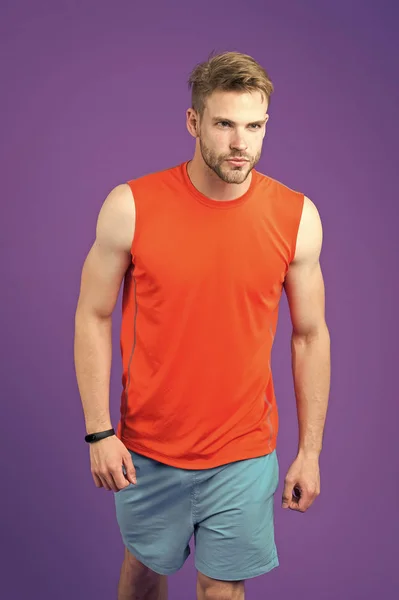 Athlete with sexy muscular body. Runner training before marathon, sport and fitness concept. Man with trimmed beard wearing sporty orange vest and blue shorts. Sportsman isolated on purple background