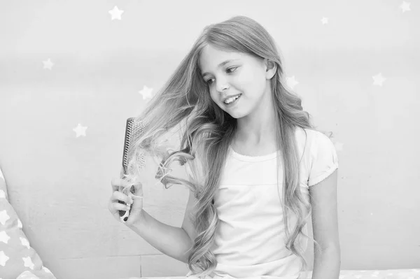 Child curly hairstyle hold hairbrush or comb. Apply oil before combing hair. Healthy hair. Conditioner or mask organic oil comb hair. Beauty salon tips. Girl long curly hair grey interior background — Stock Photo, Image