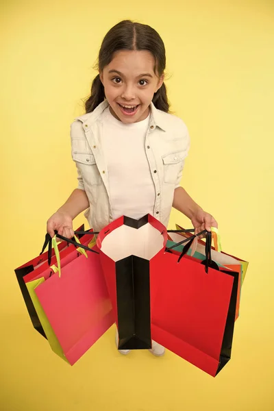 Míralo. Niño lindo fashionista compras. Niña feliz cara sonriente lleva paquetes de racimo fondo amarillo. Chica adicta a las compras le gusta ir de compras en la temporada de venta. Niño feliz compras en el centro comercial —  Fotos de Stock