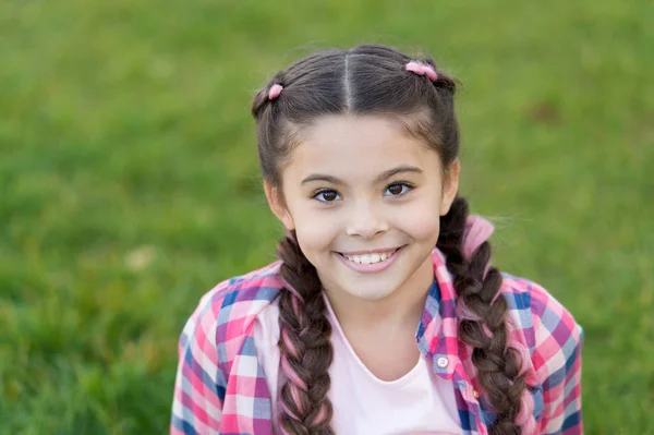 Sorriso encantador. Menina pequena criança com penteado de tranças na moda. Tendência moda. Salão e cuidados com o cabelo. Menina sorrir rosto ao ar livre. Caminhada agradável no parque. Sorriso e alegria. Penteado na moda para crianças — Fotografia de Stock