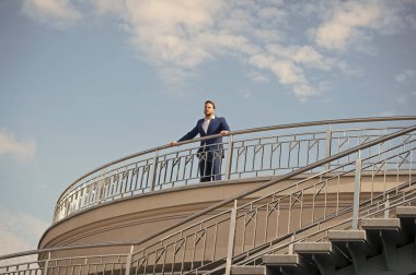 Kendine güvenen işadamı bulutlu mavi gökyüzünün balkonunda duruyor. Resmi kıyafetli adam açık havada giyiniyor. Geleceğe bakıyorum. Yeni olasılıkları düşünüyorum. Özgüven ve karizma. Erkek görünüşü. Gelmiş geçmiş en iyi yönetici.