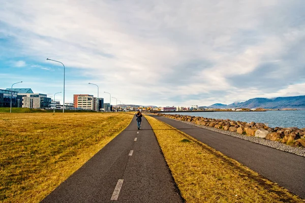 Woman walk on road along sea on cloudy sky in reykjavik, iceland. Promenade at seaside on cityscape. Travel and wanderlust on urban landscape. Freedom, perspective and future — Stock Photo, Image
