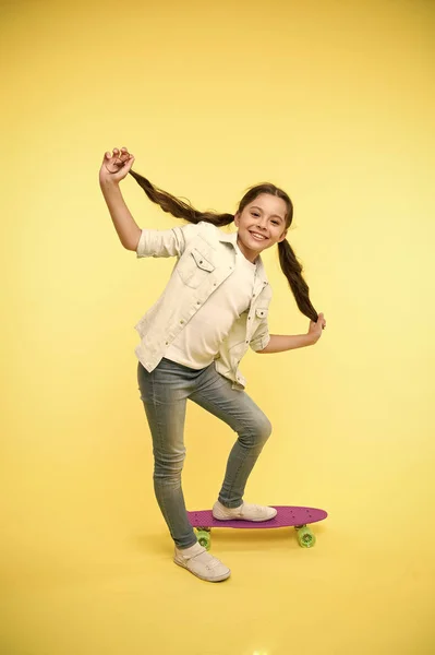 Felice ragazza hipster mostra lunghi capelli castani su penny board su sfondo giallo. Piccolo sorriso bambino con nuova acconciatura. Orgogliosa dei suoi capelli sani. Bellezza sullo skateboard. Salone di bellezza. Stile ed energia — Foto Stock