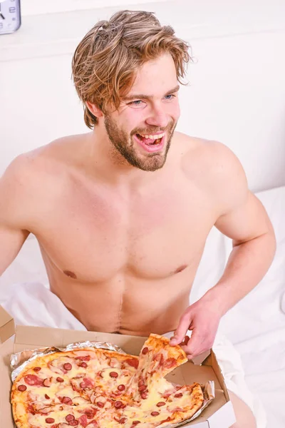 Shirtless handsome young man with pizza on bed. Guy holds pizza box sit bed in bedroom or hotel room. Portrait of lazy muscular man eating pizza while laying on a bed at home. Pizza on bed. — Stock Photo, Image