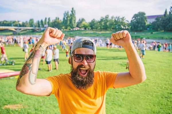 Cheerful fan at summer fest. Man bearded hipster in front of crowd people raise fists green riverside background. Urban event celebration. Hipster in cap happy celebrate event picnic fest or festival