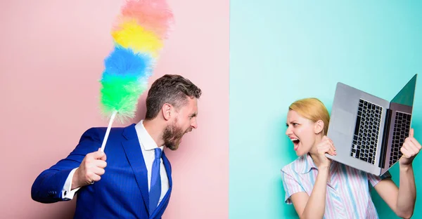 A fight between members of household. Couple of bearded man and sexy woman. Businessman and housewife. Family couple. Husband stand with dust brush while wife holding laptop. Housekeeping or business