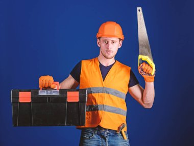 Kask, kask içinde adam araç kutusu taşır ve el testeresi, mavi arka plan tutar. Marangoz kavramı. İşçi, repairer, tamirci üzerinde ciddi bir yüz taşıyan araç, onarım için kopya alanı hazır