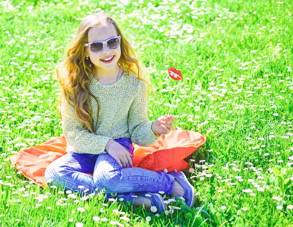 Dziewczyna siedzi na trawie o trawnik, zielone tło. Dziecko z kartonu okulary uśmiechający się warg na sesji zdjęciowej w meadow. Dziewczyna na twarz wesoły spędzić aktywny wypoczynek na świeżym powietrzu. Koncepcja mody — Zdjęcie stockowe