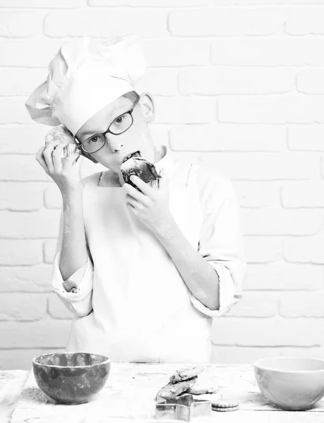 Jovem menino cozinheiro bonito chef em uniforme branco e chapéu na farinha de rosto manchado com óculos de pé perto da mesa com tigelas coloridas e segurando bolos de chocolate no fundo da parede de tijolo — Fotografia de Stock