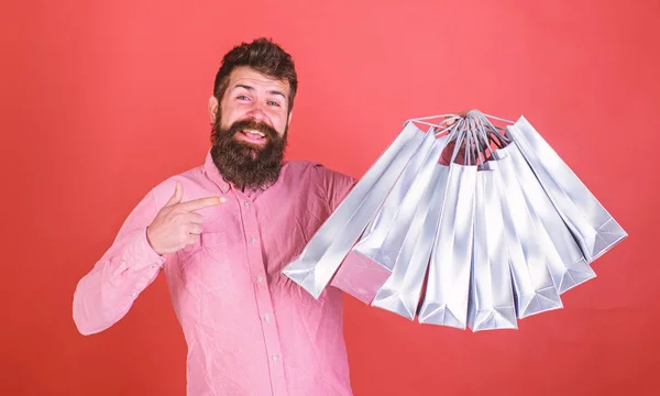 Mann mit Bart und Schnurrbart zeigt auf Einkaufstüten, roter Hintergrund. Hipster mit glücklichem Gesicht sind shoppingsüchtig oder shopaholic. Typen, die in der Verkaufssaison mit Rabatten einkaufen. Einkaufskonzept — Stockfoto