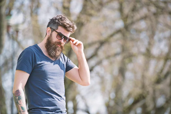 Hipster mannen i trendiga solglasögon poserar på gränden med kala träd. Skäggig man i mörk blå T-shirt med tatuering på hans arm side se i stadsbilden solig dag — Stockfoto