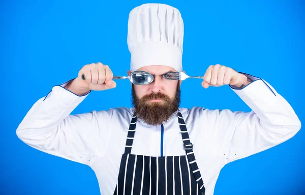 Chef sério rosto estrito segurar colher e garfo. Conceito de processo de cozinha. Vamos experimentar o prato. Chef faminto pronto para experimentar comida. Está na hora de provar. Homem bonito com barba segura utensílios de cozinha no fundo azul — Fotografia de Stock