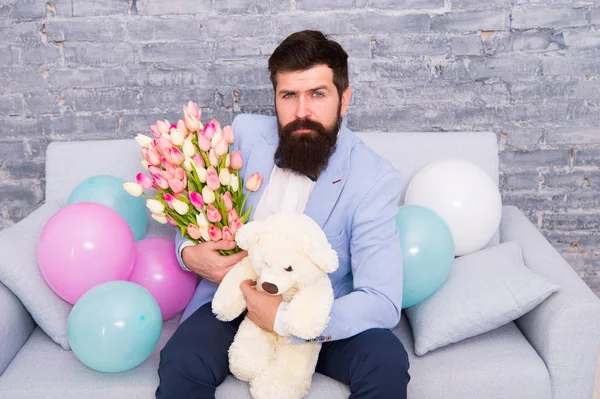 Romantic man with flowers and teddy bear sit on couch waiting girlfriend. Romantic gift. Macho getting ready romantic date. Man wear blue tuxedo bow tie hold flowers bouquet. Best boyfriend ever