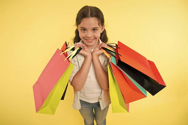 Ekspertów mody. Dziecko ładny zakupy expert pomaga wykonać pakietów podczas zakupów. Mały sklep ekspert. Shopaholic dziewczyna lubi zakupy. Dziecko dziewczynka szczęśliwy twarz prowadzi kilka pakietów żółte tło — Zdjęcie stockowe