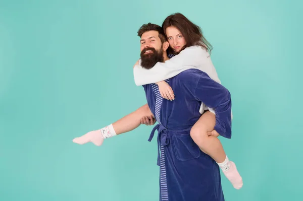 Couple in bathrobes having fun turquoise background. Lets stay at home and have fun. They always have fun together. Close relationship. Handsome young man giving his girlfriend piggyback ride