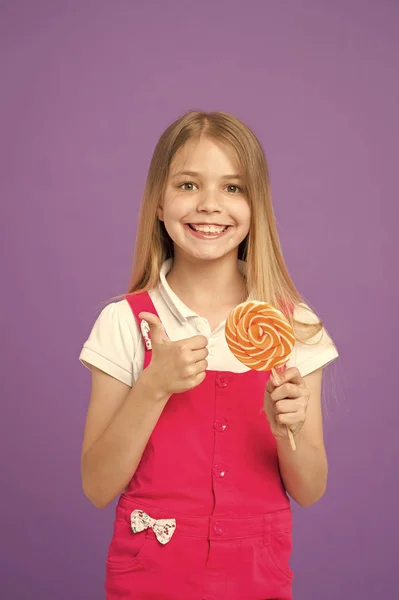 Tjej äter färgglada lollipop. Söta barndom koncept. Barn med långt hår gillar sötsaker. Flicka med leende ansikte håller jätte godis i handen, violett bakgrund. Barn i rosa overaller visar tummen — Stockfoto